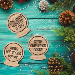 Op de afbeelding: Houten kerstversieringen met de tekst: "Give your parents a hug", "Make hot cocoa bombs" en "Send Christmas cards". De ornamenten zijn rond met een voet, geplaatst op een turquoise houten oppervlak met dennenappels en groen.