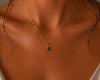 May include: A delicate gold chain necklace featuring a small, teardrop-shaped green gemstone pendant. The pendant is securely set in a simple gold prong setting, hanging centrally from the thin, polished gold metal chain against the skin.