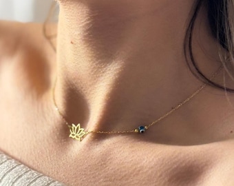 May include: Delicate gold-colored necklace featuring a lotus flower pendant and a small blue evil eye charm. The necklace is worn around the neck, showcasing its minimalist design and elegant aesthetic. The chain appears thin and refined.