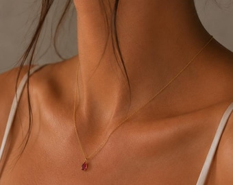 May include: A delicate gold chain necklace featuring a small, oval-shaped deep red gemstone pendant. The necklace rests against the collarbone, showcasing a thin, polished metal link chain and a single, faceted dark red stone setting.