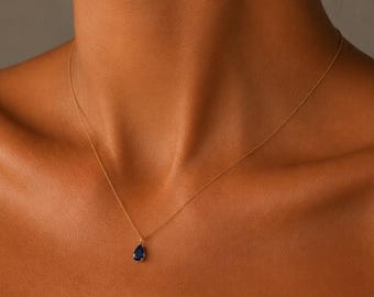 May include: A delicate gold chain necklace featuring a single teardrop-shaped dark blue gemstone pendant. The necklace rests against the collarbone, showcasing a thin metallic link chain and a polished, faceted blue stone set in a gold frame.