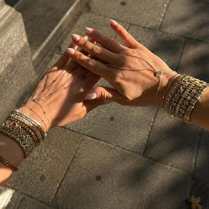 Op de afbeelding: Een collectie goudkleurige armbanden gedragen om beide polsen, met diverse schakelstijlen, pav&eacute;-gezette stenen en een enkele rode accentsteen. Een fijne gouden ketting verbindt een armband met een ring aan de middelvinger.