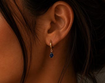 May include: A gold hoop earring featuring a small, teardrop-shaped dark blue gemstone pendant. The earring is secured through the earlobe, showcasing a polished gold metal finish and a deep blue stone set in a simple, delicate gold frame.