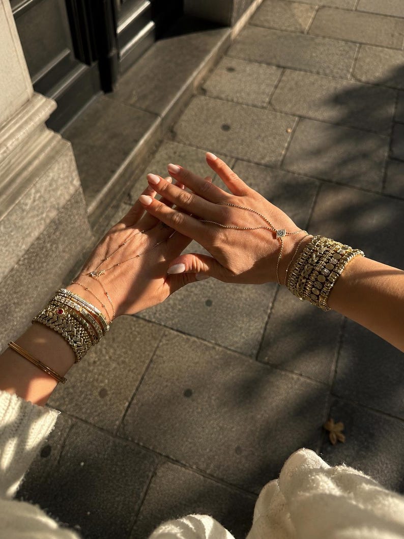 Op de afbeelding: Een collectie goudkleurige armbanden gedragen om beide polsen, met diverse schakelstijlen, pav&eacute;-gezette stenen en een enkele rode accentsteen. Een fijne gouden ketting verbindt een armband met een ring aan de middelvinger.