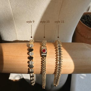 Op de afbeelding: Drie goudkleurige armbanden op een cilindrische vorm. Stijl 9 heeft afwisselend ronde en baguette-geslepen heldere stenen. Stijl 10 heeft een centrale rode ronde steen met heldere accenten. Stijl 11 bestaat uit een rij baguette-geslepen stenen.
