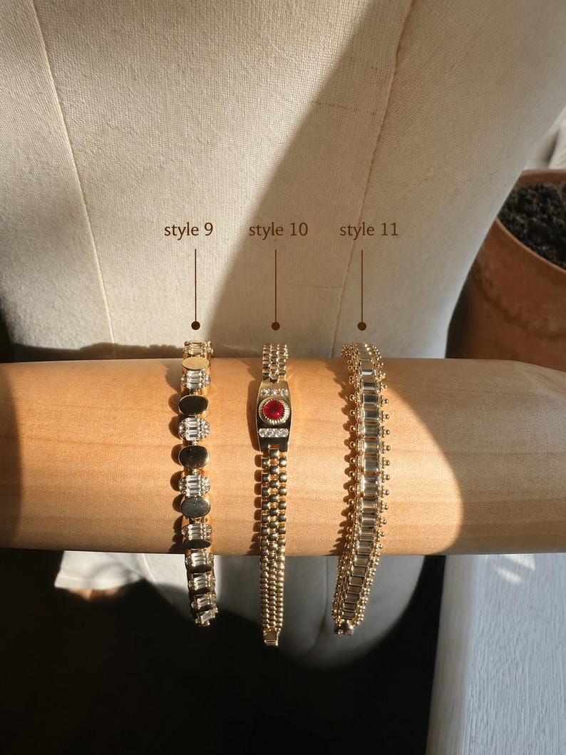 Op de afbeelding: Drie goudkleurige armbanden op een cilindrische vorm. Stijl 9 heeft afwisselend ronde en baguette-geslepen heldere stenen. Stijl 10 heeft een centrale rode ronde steen met heldere accenten. Stijl 11 bestaat uit een rij baguette-geslepen stenen.