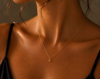 May include: A delicate gold chain necklace featuring a small, teardrop-shaped pendant. The necklace rests against the collarbone, showcasing a thin, polished metal link chain and a single, subtle gemstone charm that hangs centered at the base of the neck.