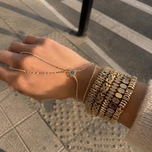 May include: A gold hand chain featuring a central evil eye charm with blue and white stones, connected to a delicate chain. The wrist is adorned with a stack of five gold bracelets, including link chains, pavé-set stones, and oval accents.