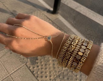 May include: A gold hand chain featuring a central evil eye charm with blue and white stones, connected to a delicate chain. The wrist is adorned with a stack of five gold bracelets, including link chains, pavé-set stones, and oval accents.
