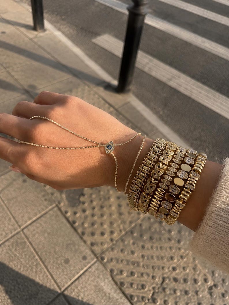 Op de afbeelding: Een gouden handketting met een centraal oog-symbool voorzien van blauwe en witte stenen, verbonden aan een fijne ketting. De pols is versierd met een stapel van vijf gouden armbanden, waaronder schakelkettingen, pav&eacute;-gezette stenen en ovale accenten.