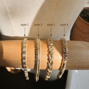 Op de afbeelding: Vier goudkleurige armbanden gepresenteerd op een cilindrische vorm. Stijl 1 heeft vierkante stenen, stijl 2 een verticaal baguette-ontwerp, stijl 3 een gevlochten schakelketting met pav&eacute;-stenen en stijl 4 bestaat uit gepolijste gouden schakels.