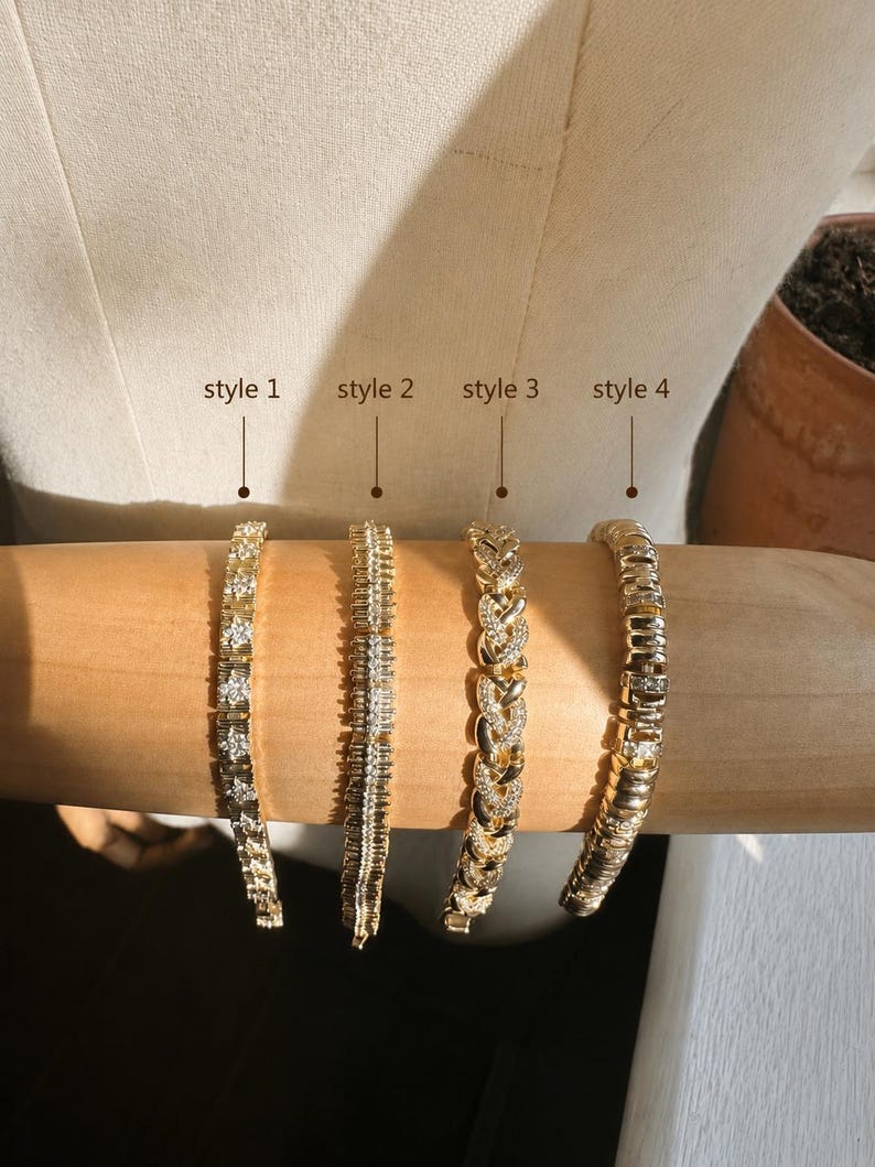 Op de afbeelding: Vier goudkleurige armbanden gepresenteerd op een cilindrische vorm. Stijl 1 heeft vierkante stenen, stijl 2 een verticaal baguette-ontwerp, stijl 3 een gevlochten schakelketting met pav&eacute;-stenen en stijl 4 bestaat uit gepolijste gouden schakels.
