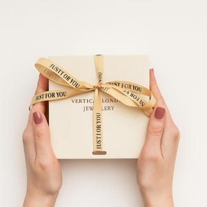 Op de afbeelding: Een vierkante off-white geschenkdoos met een gouden lint gestrikt tot een strik. Het lint is bedrukt met de zwarte tekst JUSTT OR YOU. Het deksel van de doos toont de tekst VERTICAL LOND JEWLERY in donkere letters onder de strik.