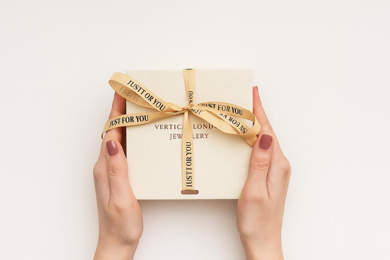 Op de afbeelding: Een vierkante off-white geschenkdoos met een gouden lint gestrikt tot een strik. Het lint is bedrukt met de zwarte tekst JUSTT OR YOU. Het deksel van de doos toont de tekst VERTICAL LOND JEWLERY in donkere letters onder de strik.