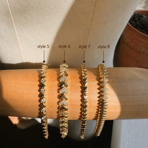 Op de afbeelding: Vier goudkleurige armbanden op een cilindrische vorm. Stijl 5 heeft hartvormige schakels met heldere stenen. Stijl 6 heeft chevronvormige schakels met pav&eacute;-stenen. Stijl 7 bestaat uit effen hartschakels. Stijl 8 heeft marquisevormige schakels.
