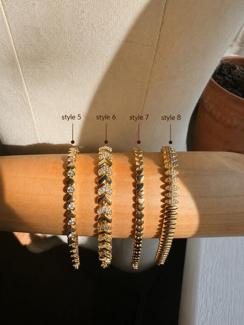 Op de afbeelding: Vier goudkleurige armbanden op een cilindrische vorm. Stijl 5 heeft hartvormige schakels met heldere stenen. Stijl 6 heeft chevronvormige schakels met pav&eacute;-stenen. Stijl 7 bestaat uit effen hartschakels. Stijl 8 heeft marquisevormige schakels.