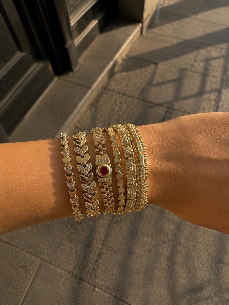 Op de afbeelding: Een stapel van vijf goudkleurige armbanden. De set bevat een armband met hartvormige schakels en heldere stenen, een armband met chevronpatroon en heldere stenen, een horloge-stijl armband met een centrale rode steen, een ketting met hartschakels en twee getextureerde armbanden met heldere stenen.