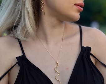 May include: Gold-colored jewelry set featuring a snake design. The set includes a necklace and earrings. The necklace hangs down the chest, and the earrings dangle from the ear. The jewelry is worn with a black top.
