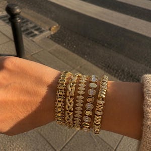 Op de afbeelding: Een stapel van vijf goudkleurige armbanden met gevarieerde ontwerpen, waaronder een schakelketting, een gevlochten patroon, een zetting met marquisevormige stenen, een reeks ovale stenen en een getextureerde band met heldere kristallen.
