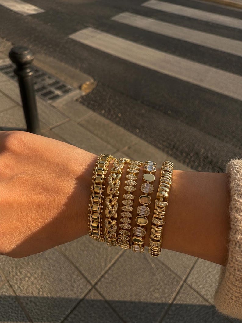 Op de afbeelding: Een stapel van vijf goudkleurige armbanden met gevarieerde ontwerpen, waaronder een schakelketting, een gevlochten patroon, een zetting met marquisevormige stenen, een reeks ovale stenen en een getextureerde band met heldere kristallen.