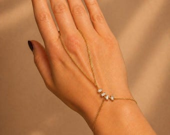 May include: A delicate gold hand chain featuring three clear, rectangular gemstones. The chain connects to the wrist and finger, creating a minimalist and elegant jewelry piece. The hand chain is displayed against a neutral background.
