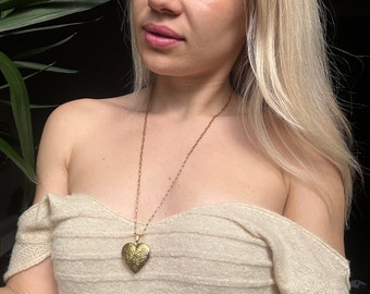 May include: A gold heart-shaped locket necklace with an intricate design. The necklace is worn by a person with long blonde hair and a white top.