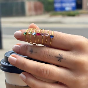 May include: A selection of gold-coloured rings, each with a different coloured oval gemstone. Gemstones include green, clear, light blue, red, purple, pink, and blue. The rings are displayed on a finger.