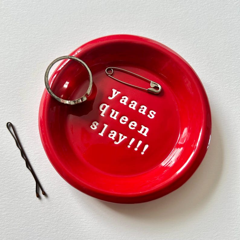 May include: A bright red ceramic trinket dish with the text "Yaaas Queen Slay!!!" in white. A silver ring, a safety pin, and a black bobby pin are inside the dish. The dish is round and sits on a white surface.