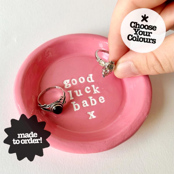 May include: A round pink ceramic trinket dish featuring the white embossed text "good luck babe x" in the center. Two silver rings are placed inside the dish. A white circular sticker with the text "Choose Your Colours" is visible.