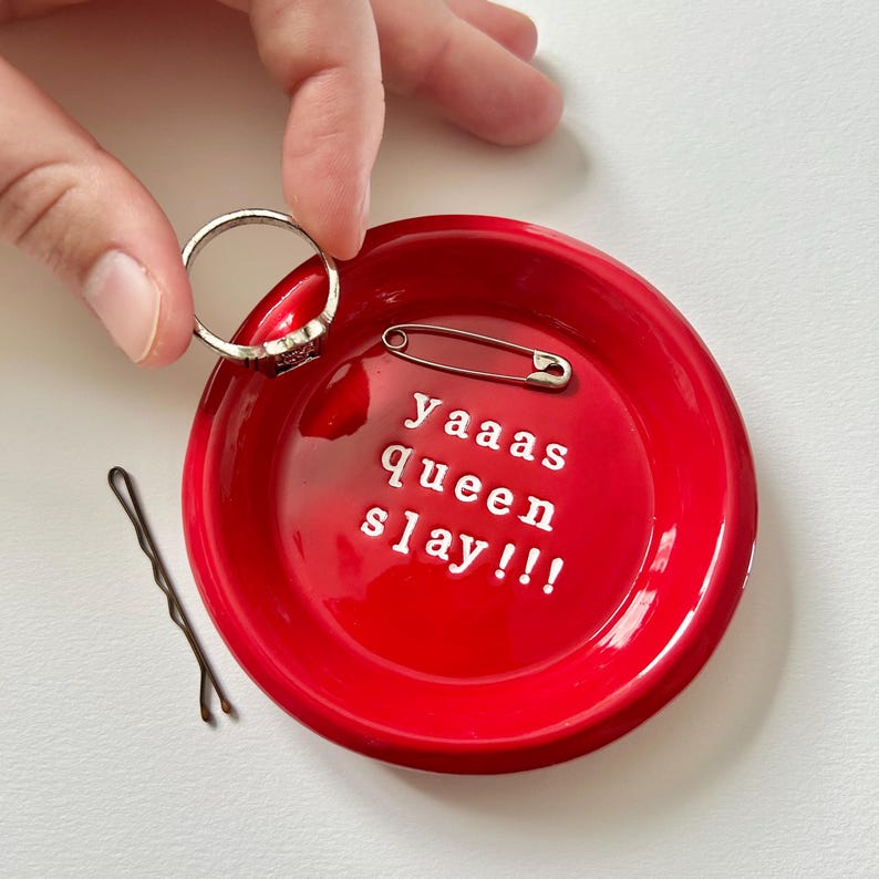 May include: A bright red trinket dish with the words "Yaaas Queen Slay!!!" in white lettering. A silver ring and safety pin rest inside the dish, with a bobby pin to the side. A hand is holding the ring.