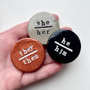 May include: Three circular pronoun badges. One is copper-coloured with "they/them" in white. Another is grey with "she/her" in white. The third is black with "he/him" in white. The badges are held in a hand.