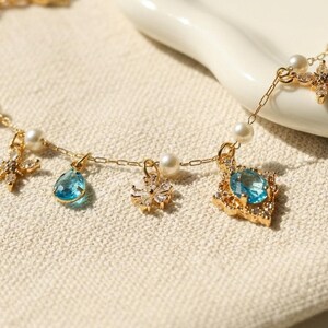 May include: A delicate gold necklace featuring various charms, including a teardrop-shaped blue gemstone, a snowflake, and a square-cut blue gemstone. The necklace also includes small pearls and star-shaped accents.