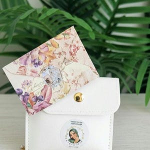 May include: A white leatherette pouch with a gold snap closure, holding a floral patterned envelope. The envelope features purple grapes and flowers. A sticker with a woman's portrait is on the pouch. A gold chain is visible.