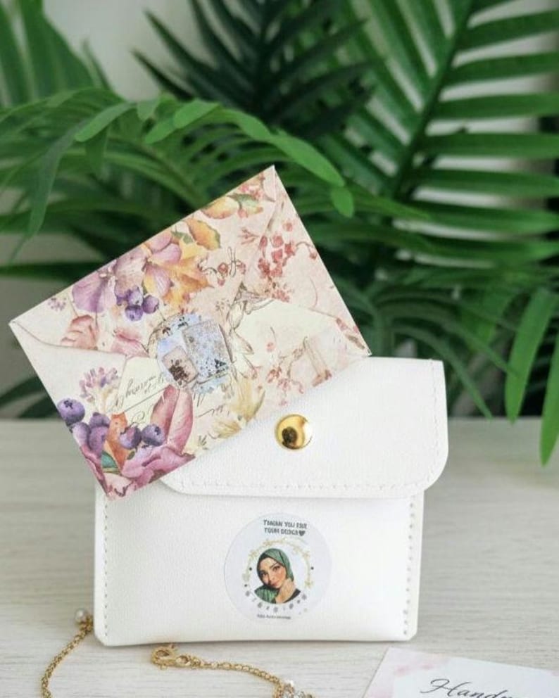 May include: A white leatherette pouch with a gold snap closure, holding a floral patterned envelope. The envelope features purple grapes and flowers. A sticker with a woman's portrait is on the pouch. A gold chain is visible.