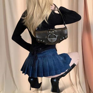 Puede incluir: Una mujer lleva una camisa negra de manga larga y una minifalda de mezclilla con pliegues. Lleva un bolso de mano de cuero marrón con herrajes plateados y tachuelas.
