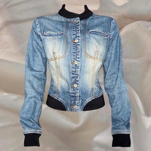 Könnte beinhalten: Hellblaue Jeansjacke mit taillierter Silhouette. Die Jacke hat einen schwarzen Kragen, Manschetten und Saum, eine Knopfleiste und zwei kleine Brusttaschen. Das Design umfasst dekorative Nähte und eine verwaschene Optik.