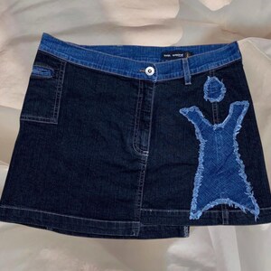Könnte beinhalten: Ein dunkelblauer Jeansrock mit einem kontrastierenden blauen Jeans-Patch in Form einer Person. Der Rock hat einen Reißverschluss und einen Knopfverschluss.