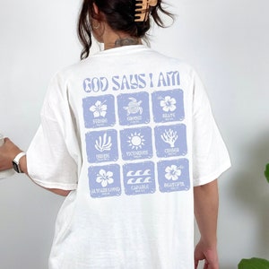 May include: White t-shirt with a blue graphic print that says "God Says I Am" and includes nine squares with different images and words, including "Strong", "Composed", "Brave", "Loving", "Victorious", "Courage", "Always Loved", "Capable", and "Beautiful".