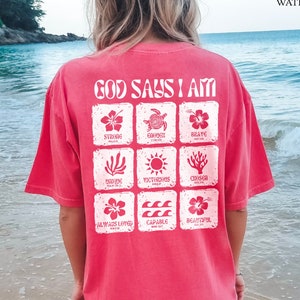 May include: A pink t-shirt with a white graphic that says "God Says I Am" and features a grid of nine squares with different images and words, including "Strong", "Brave", "Victorious", "Chosen", "Always Loved", and "Capable".