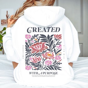 Puede incluir: Sudadera blanca con capucha con un diseño floral y el texto "CREATED WITH A PURPOSE" y una referencia bíblica "EPHESIANS 2:10 NIV".