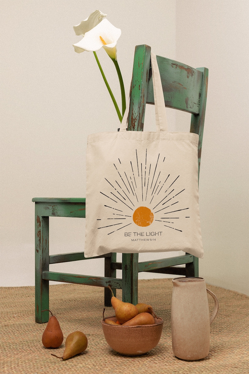 Op de afbeelding: Cr&egrave;mekleurige draagtas met een zonnestraalontwerp en de tekst "BE THE LIGHT". De tas hangt aan een verweerde groene stoel. Peren en een kan staan op de voorgrond.