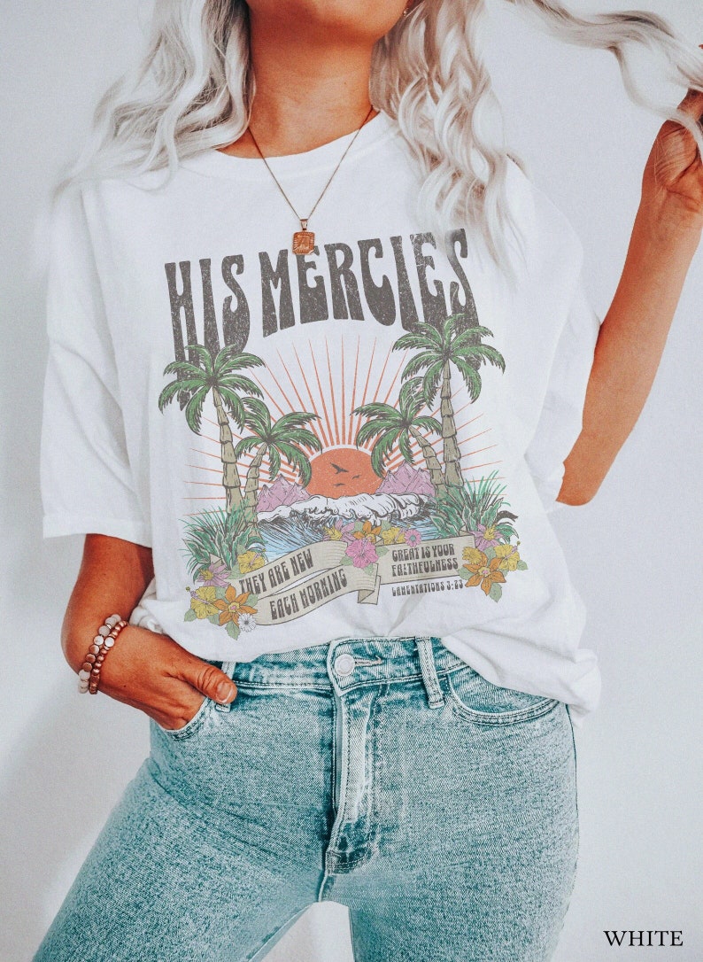 May include: White t-shirt with a graphic print of palm trees, a sunset, and the text "His Mercies" in a retro font. The text "They are new each morning" and "Great is your faithfulness" are also printed on the shirt.