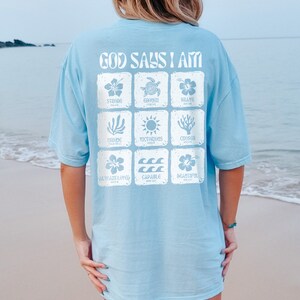 May include: A light blue t-shirt with the text "God Says I Am" printed on the back. The text is surrounded by nine squares, each containing a different image and word, such as "Strong", "Enough", "Brave", "Victorious", "Chosen", "Always Loved", "Capable", and "Beautiful".