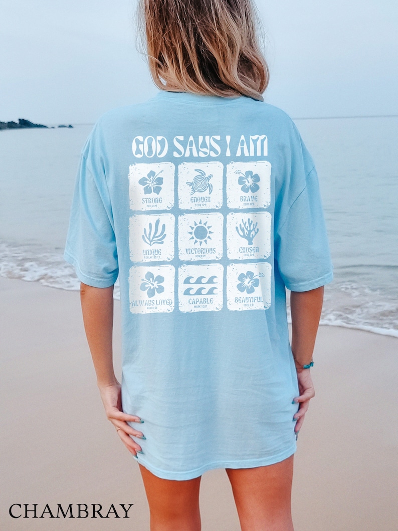 May include: A light blue t-shirt with the text "God Says I Am" printed on the back. The text is surrounded by nine squares, each containing a different image and word, such as "Strong", "Enough", "Brave", "Victorious", "Chosen", "Always Loved", "Capable", and "Beautiful".