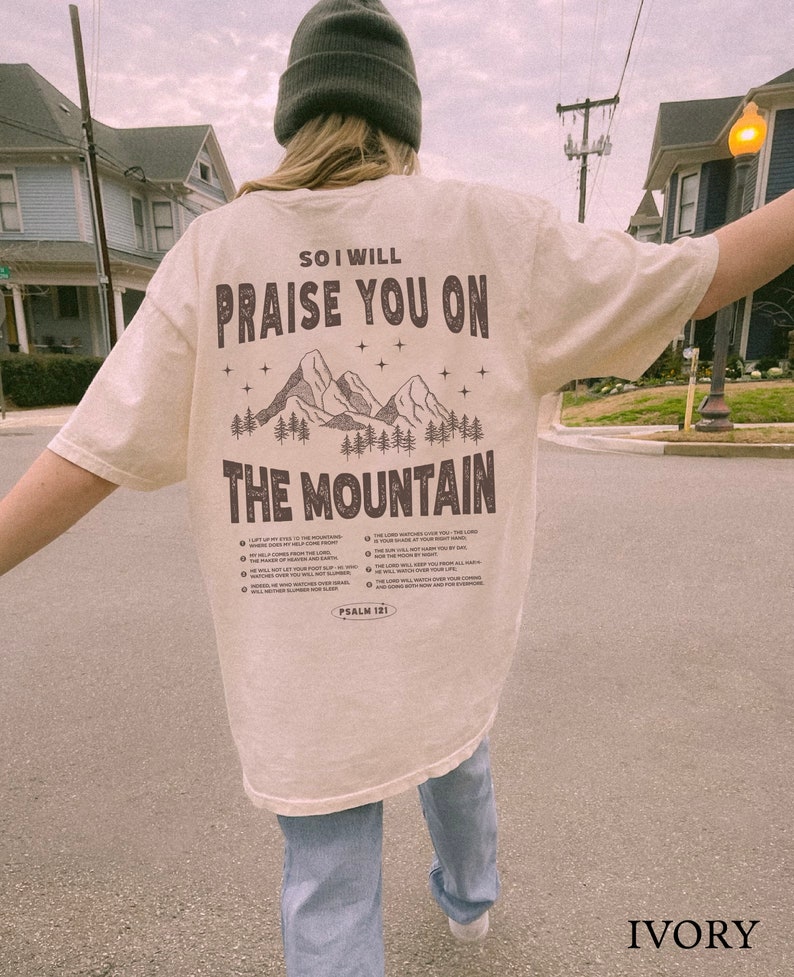 May include: Ivory-colored t-shirt with the text "So I will praise you on the mountain" in brown. The design includes mountain and tree graphics. The text "Psalm 121" is at the bottom. The shirt is worn by a person.