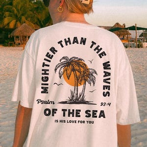 Puede incluir: Camiseta blanca con un gr&aacute;fico negro de palmeras y un sol. El texto "MIGHTER THAN THE WAVES" est&aacute; en un c&iacute;rculo alrededor del gr&aacute;fico. El texto "Psalm 93:4" est&aacute; debajo del gr&aacute;fico. El texto "OF THE SEA IS HIS LOVE FOR YOU" est&aacute; debajo del texto del Salmo.