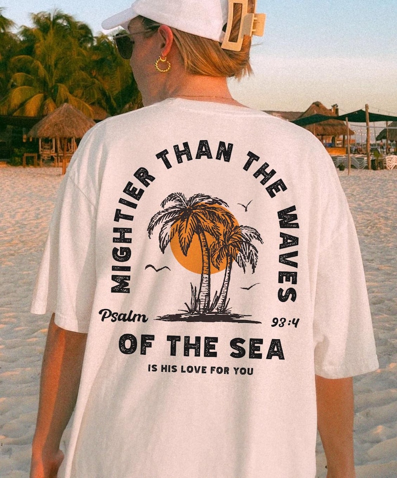 Puede incluir: Camiseta blanca con un gr&aacute;fico negro de palmeras y un sol. El texto "MIGHTER THAN THE WAVES" est&aacute; en un c&iacute;rculo alrededor del gr&aacute;fico. El texto "Psalm 93:4" est&aacute; debajo del gr&aacute;fico. El texto "OF THE SEA IS HIS LOVE FOR YOU" est&aacute; debajo del texto del Salmo.