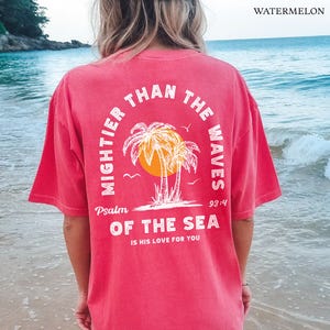 Puede incluir: Una camiseta roja con un dise&ntilde;o gr&aacute;fico blanco con palmeras y el texto "Mightier than the waves Psalm 93:4 of the sea is his love for you".