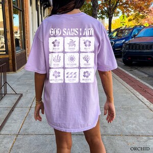 May include: A lavender-colored t-shirt with the text "God Says I Am" printed on the back. The shirt features a grid of nine squares, each containing a different image and word, such as "Strong", "Brave", "Capable", and "Beautiful".