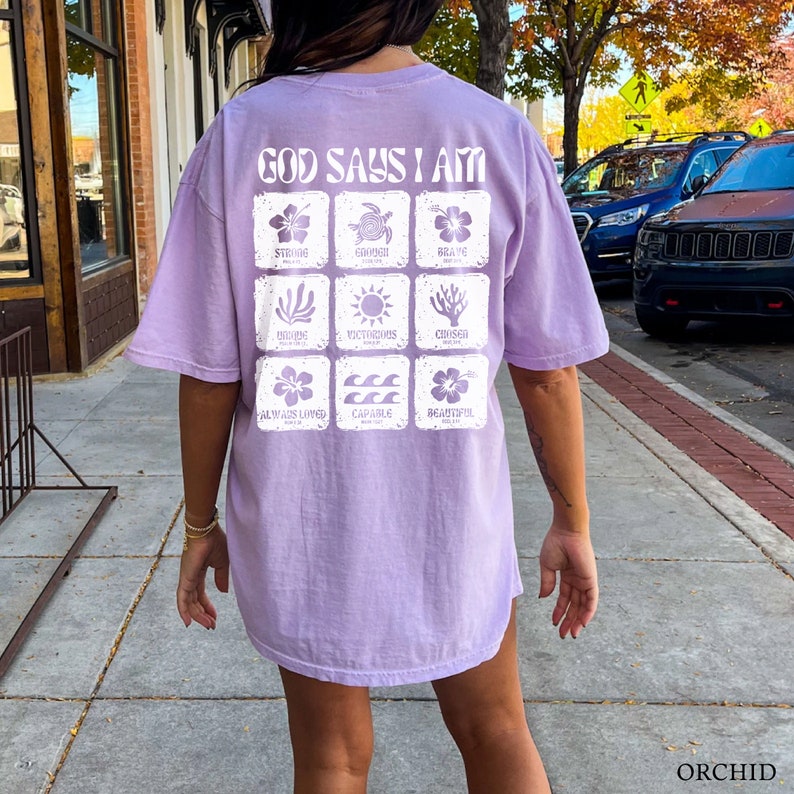 May include: A lavender-colored t-shirt with the text "God Says I Am" printed on the back. The shirt features a grid of nine squares, each containing a different image and word, such as "Strong", "Brave", "Capable", and "Beautiful".